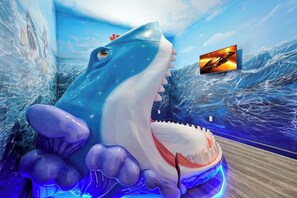 A large shark sculpture in a room with a wall mural of ocean waves, a television screen, and a wooden floor.