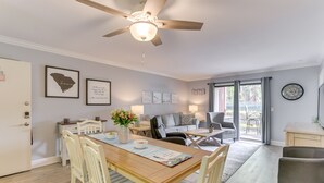 A dining area with a wooden table, chairs, and a ceiling fan. There are framed pictures on the wall and a window with curtains.