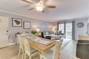 A dining area with a wooden table, chairs, and a ceiling fan. There are framed pictures on the wall and a window with curtains.