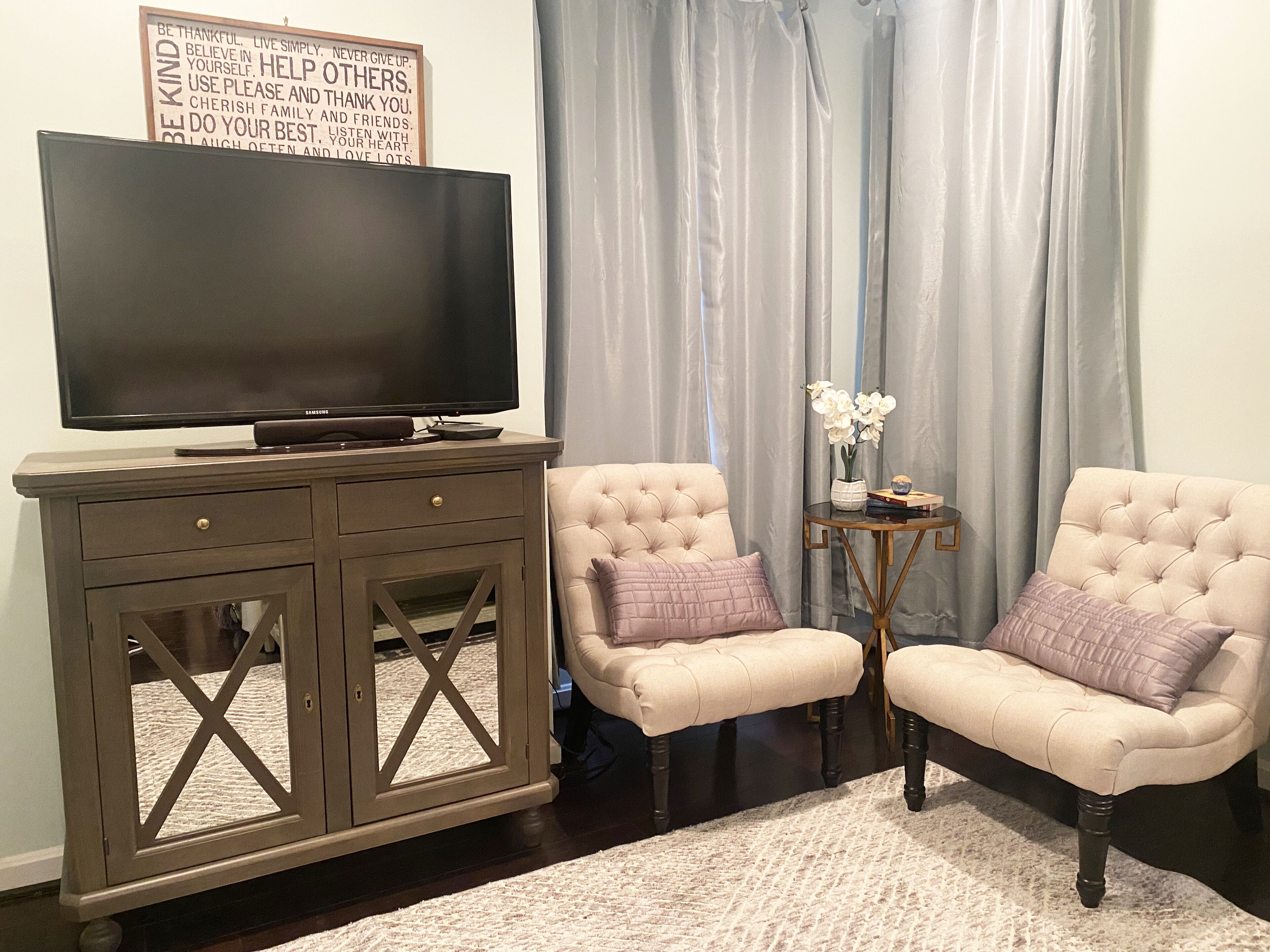 A living room with a television on a cabinet, two armchairs, a small table with a vase of flowers, and a sign with inspirational quotes.