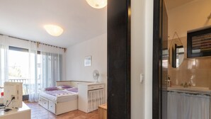 A bedroom with a bed, bedside table, and a desk with a computer. There is a window with curtains and a view of the outside.