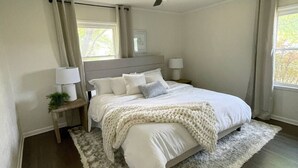 Une chambre à coucher avec un grand lit, deux tables de chevet avec des lampes, un ventilateur de plafond et une vue sur les arbres par la fenêtre.