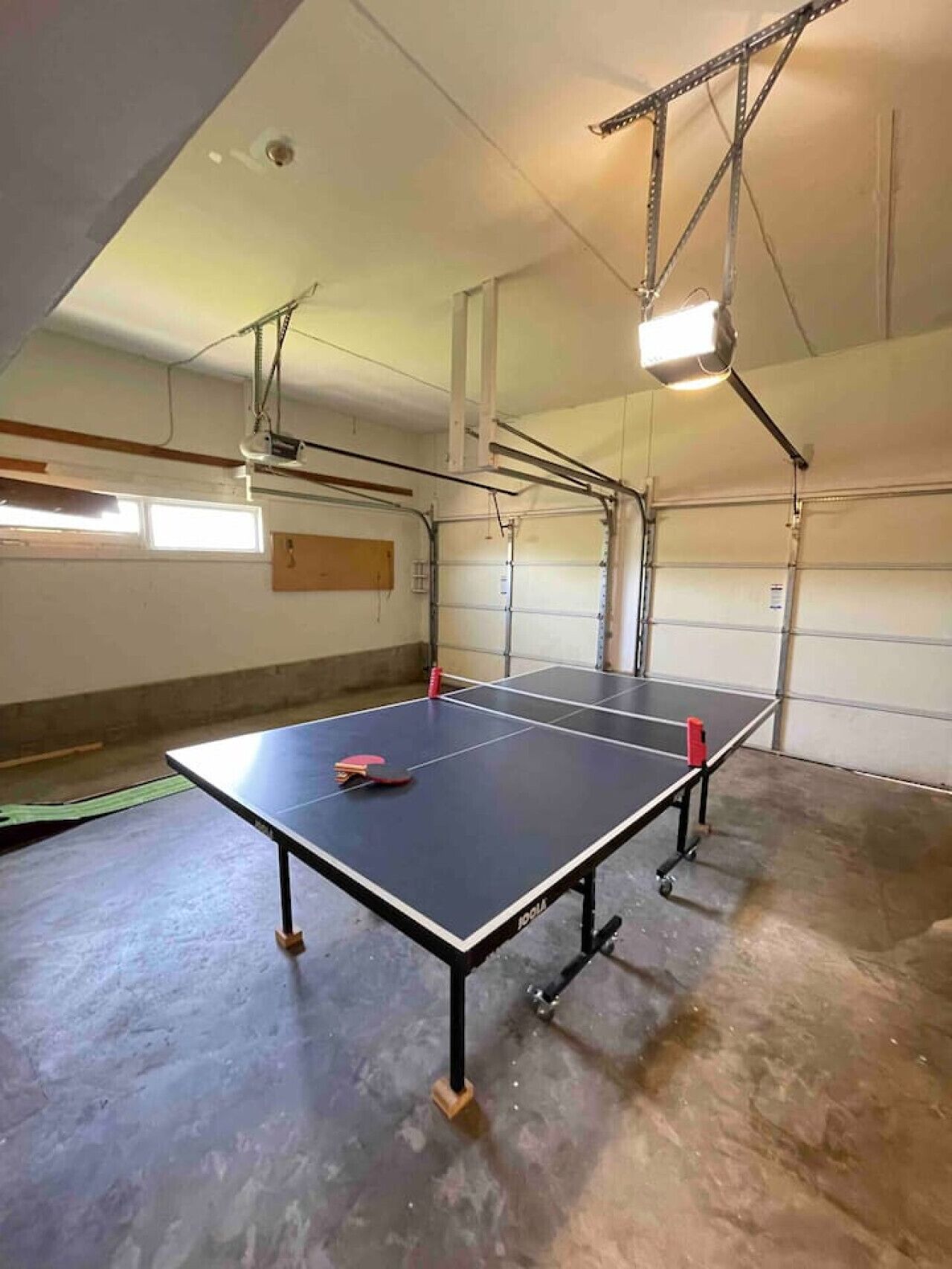 A spacious indoor area with two ping pong tables, a ceiling-mounted projector, and a garage door in the background.