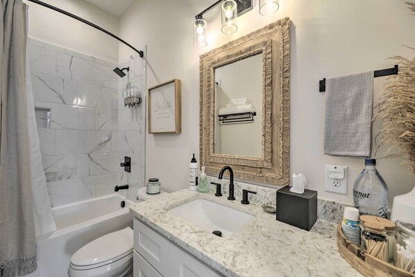 A modern bathroom with a bathtub, a sink, and a large mirror. There are toiletries on the counter and a towel rack above the mirror.