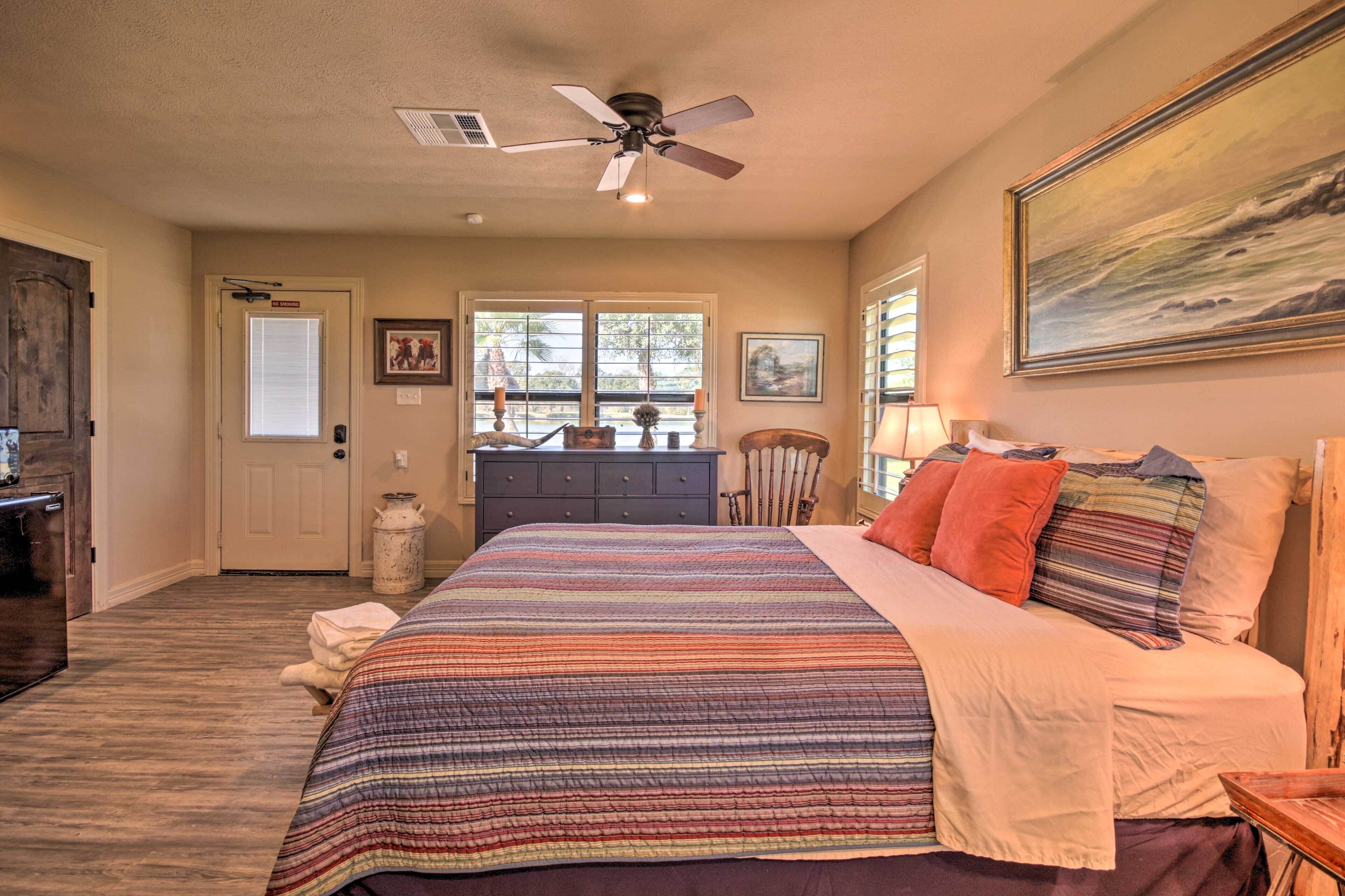 A bedroom with a large bed, a ceiling fan, a door with a window, and a painting on the wall.