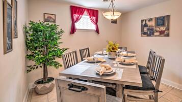 A dining area with a wooden table set for six, surrounded by chairs, a potted plant, and a window with red curtains.