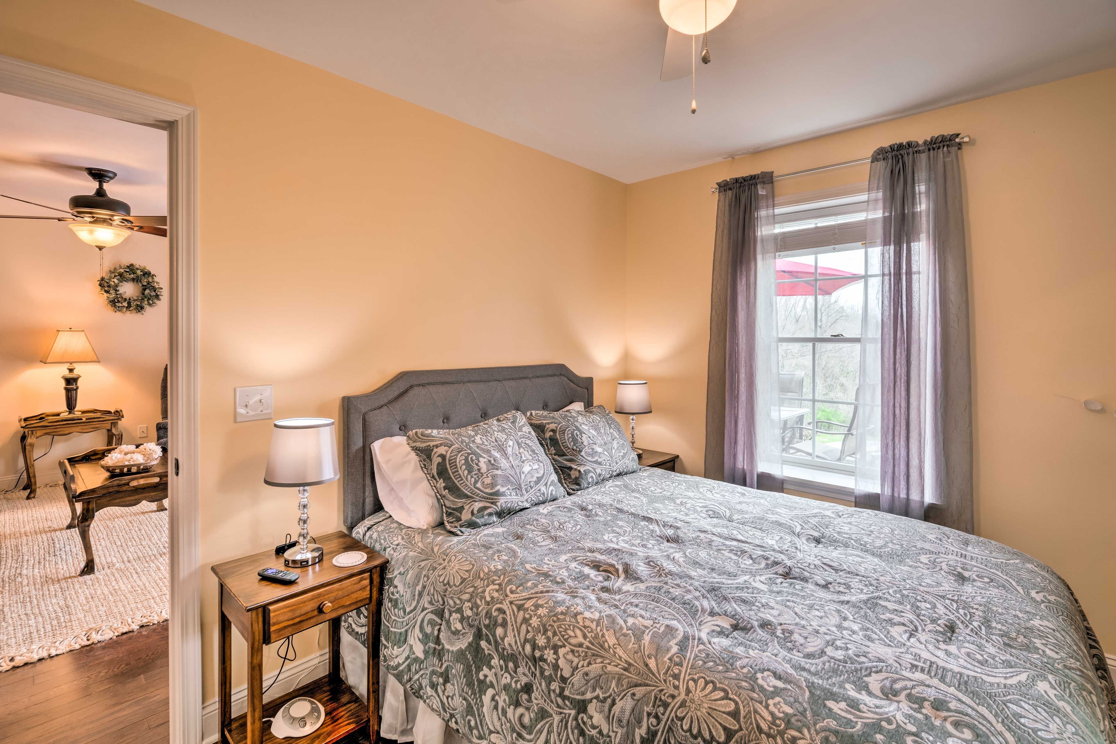 Een slaapkamer met een bed, nachtkastjes, een lamp, een plafondventilator, een raam met gordijnen en uitzicht op de omgeving.
