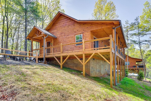 A wooden cabin with a deck and a covered porch, surrounded by trees.