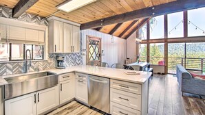 Una cocina moderna con armarios blancos, electrodomésticos de acero inoxidable y una isla grande. Hay un comedor con una mesa y sillas, y vistas al exterior a través de amplias ventanas.
