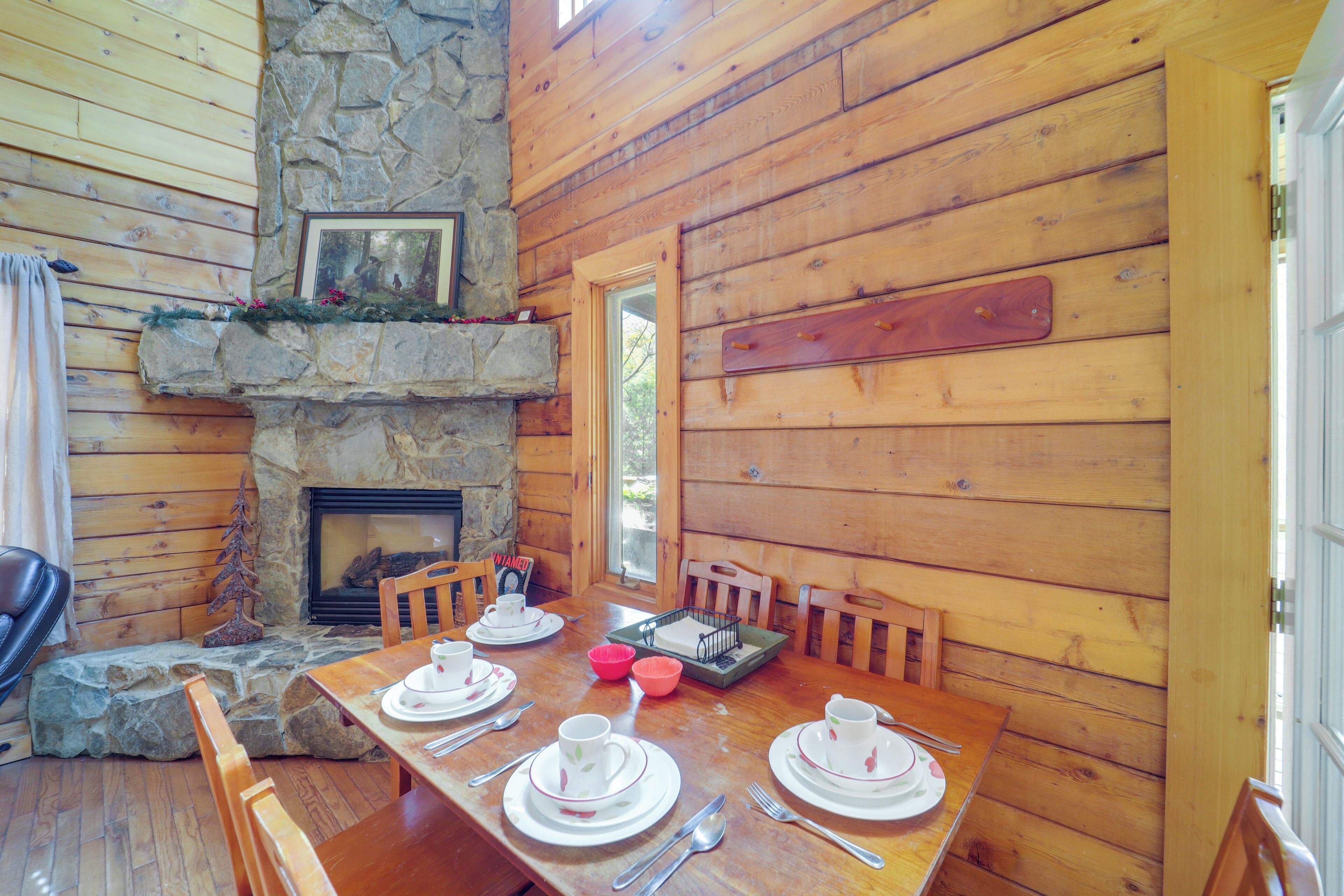 A wooden dining table set with white plates, cups, and cutlery, surrounded by wooden chairs, with a stone fireplace and wooden walls.