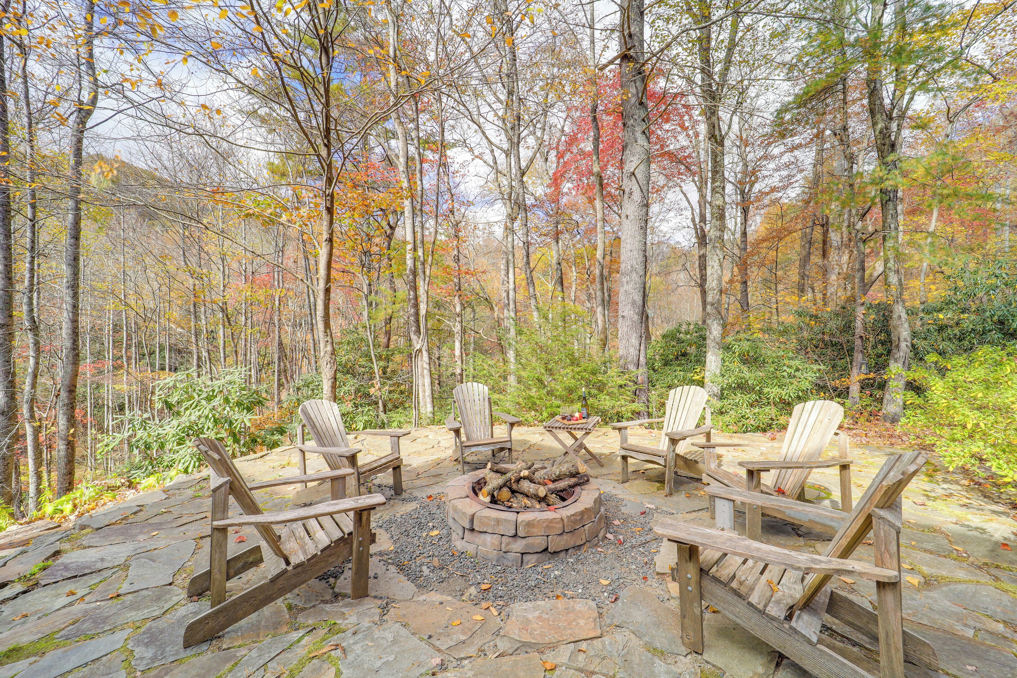 A circular stone fire pit surrounded by wooden Adirondack chairs in a forest setting.