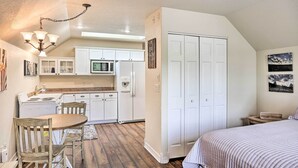 A compact kitchen with white cabinets, a microwave, and a refrigerator. There is a dining table with chairs and a chandelier. A bed with a striped blanket is visible in the foreground. - Central Creekfront Spearfish Apt by City Park (Spearfish)