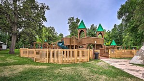 A playground with a wooden play structure, including a slide and climbing areas, surrounded by a wooden fence and trees. - Central Creekfront Spearfish Apt by City Park (Spearfish)