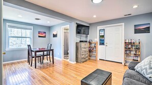 A spacious living room with hardwood floors, a dining area with a table and chairs, a television mounted on the wall, a bookshelf, and a door leading to another room.
