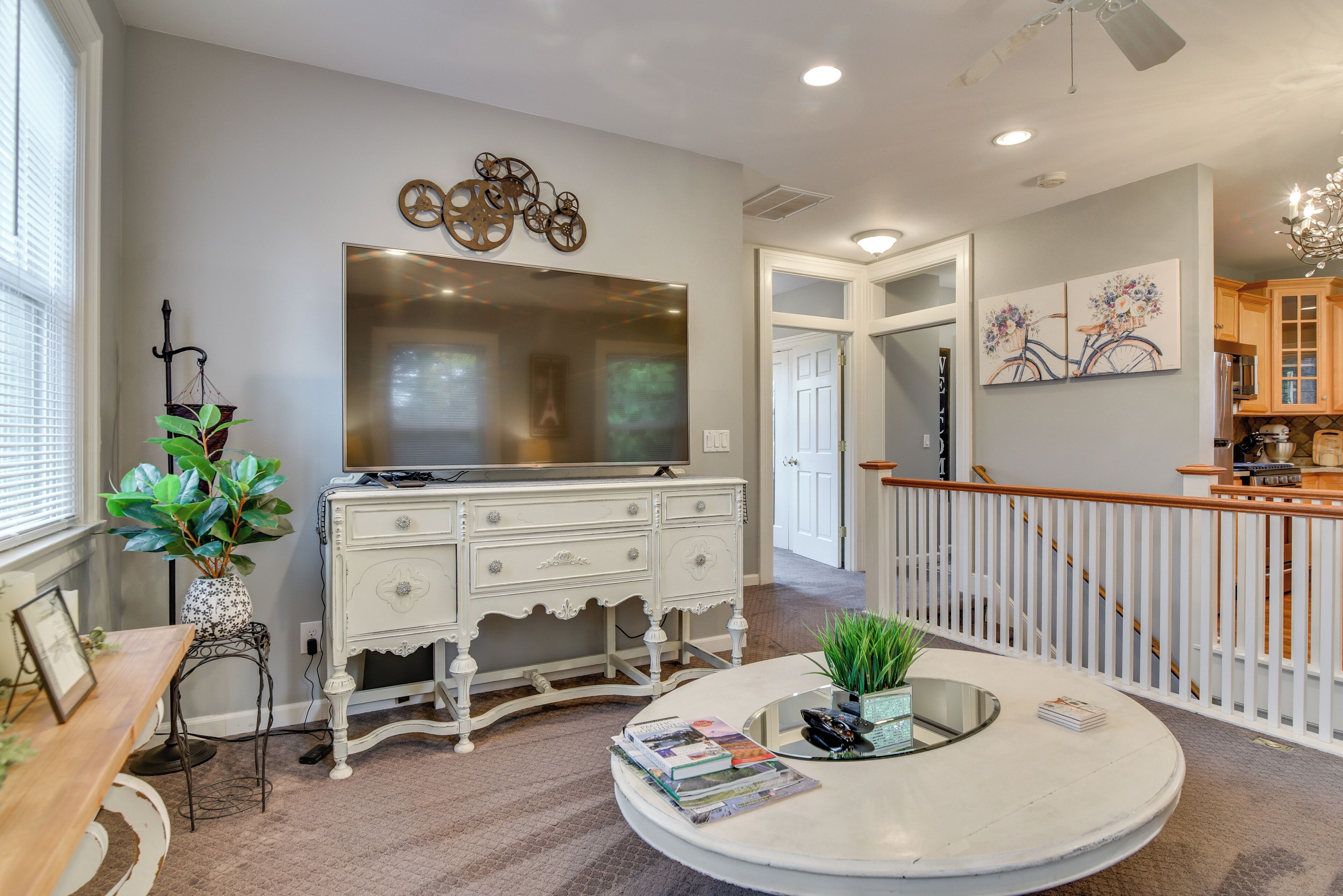 A spacious living area with a large flat-screen TV, a white ornate dresser, a round coffee table with magazines, and a staircase leading to a balcony.