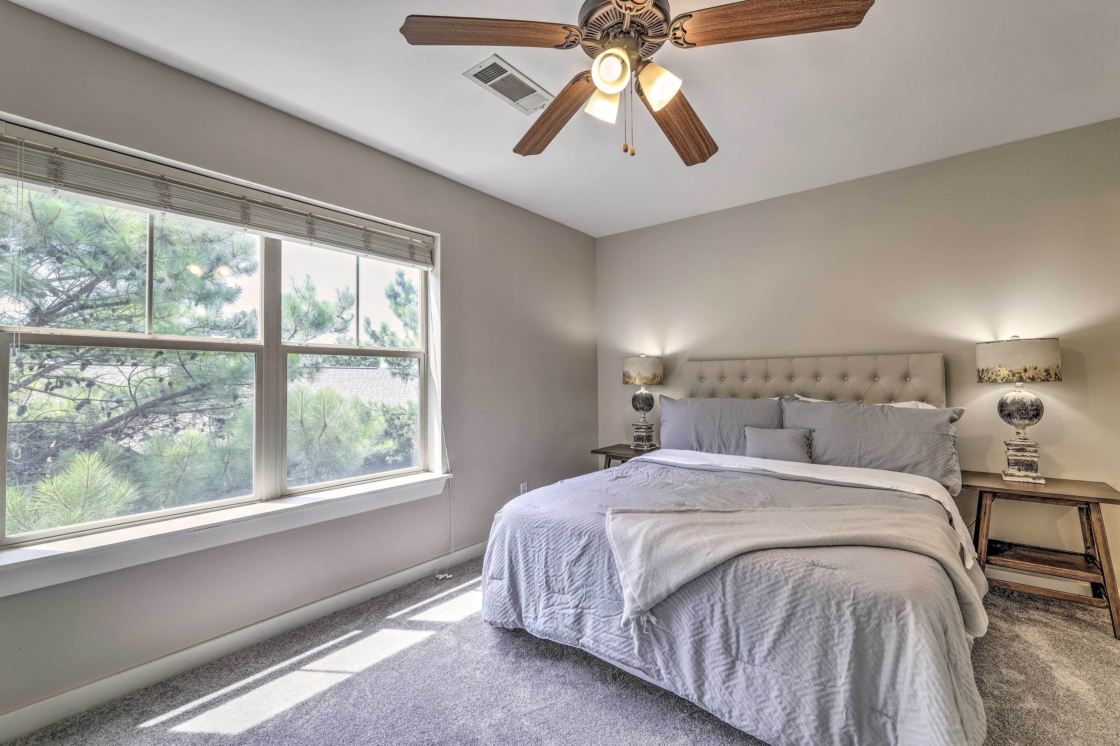 A bedroom with a large window, a bed, a ceiling fan, and a nightstand with a lamp.
