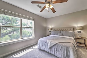 Une chambre avec une grande fenêtre, un lit, un ventilateur de plafond et une table de chevet avec une lampe.