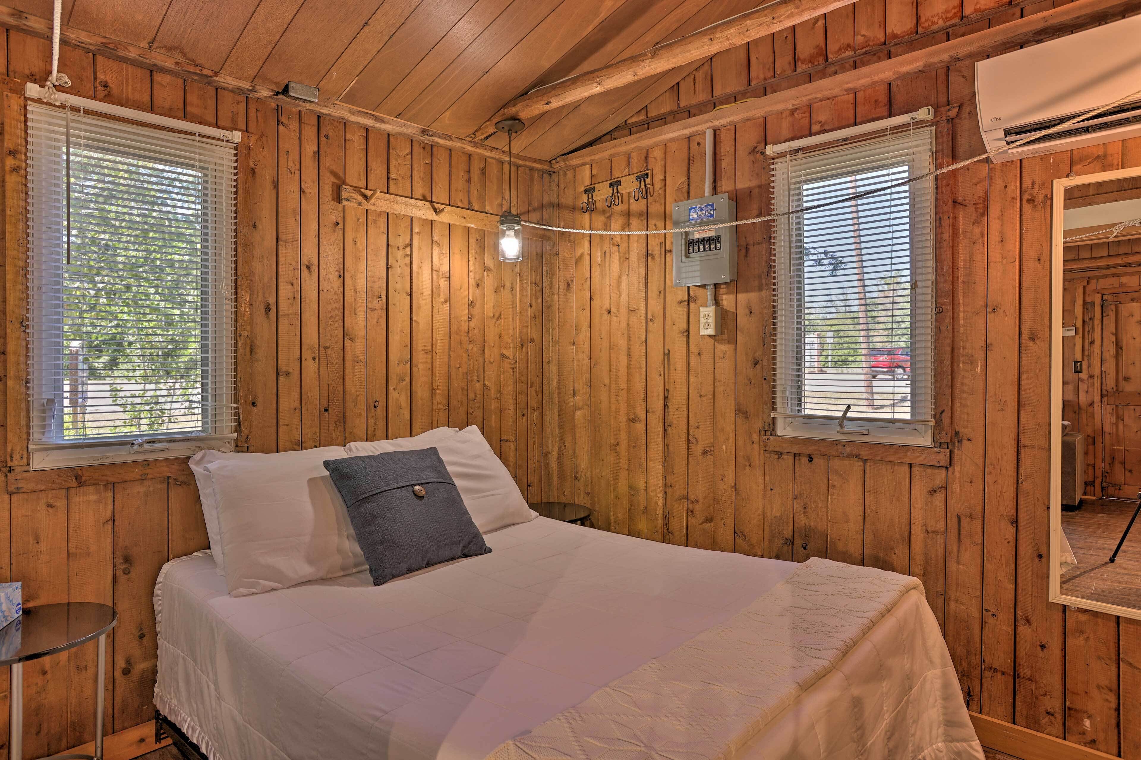 Una habitación con paneles de madera, una cama, dos ventanas con persianas, un equipo de aire acondicionado y una mesita.