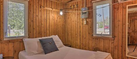 Una habitación con paneles de madera, una cama, dos ventanas con persianas, un equipo de aire acondicionado y una mesita.