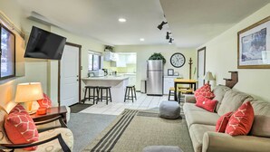 Ein modernes Wohnzimmer mit einer Couch, einem Hocker und einem Flachbildfernseher. Im Hintergrund ist eine Küche mit weißen Schränken und einem Kühlschrank zu sehen.