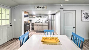 Uma sala de jantar com uma mesa branca, cadeiras azuis e uma bandeja decorativa amarela. Uma cozinha moderna visível ao fundo, com eletrodomésticos.