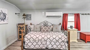Quarto com cama, mesa de cabeceira, luminária, ar-condicionado fixado na parede e janela com cortinas.