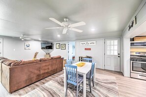A living room with a brown sofa, a dining table with blue chairs, a ceiling fan, and a kitchen area with a stove. - Cottage With Patio & Shared Waterfront Perks! (New Bern)