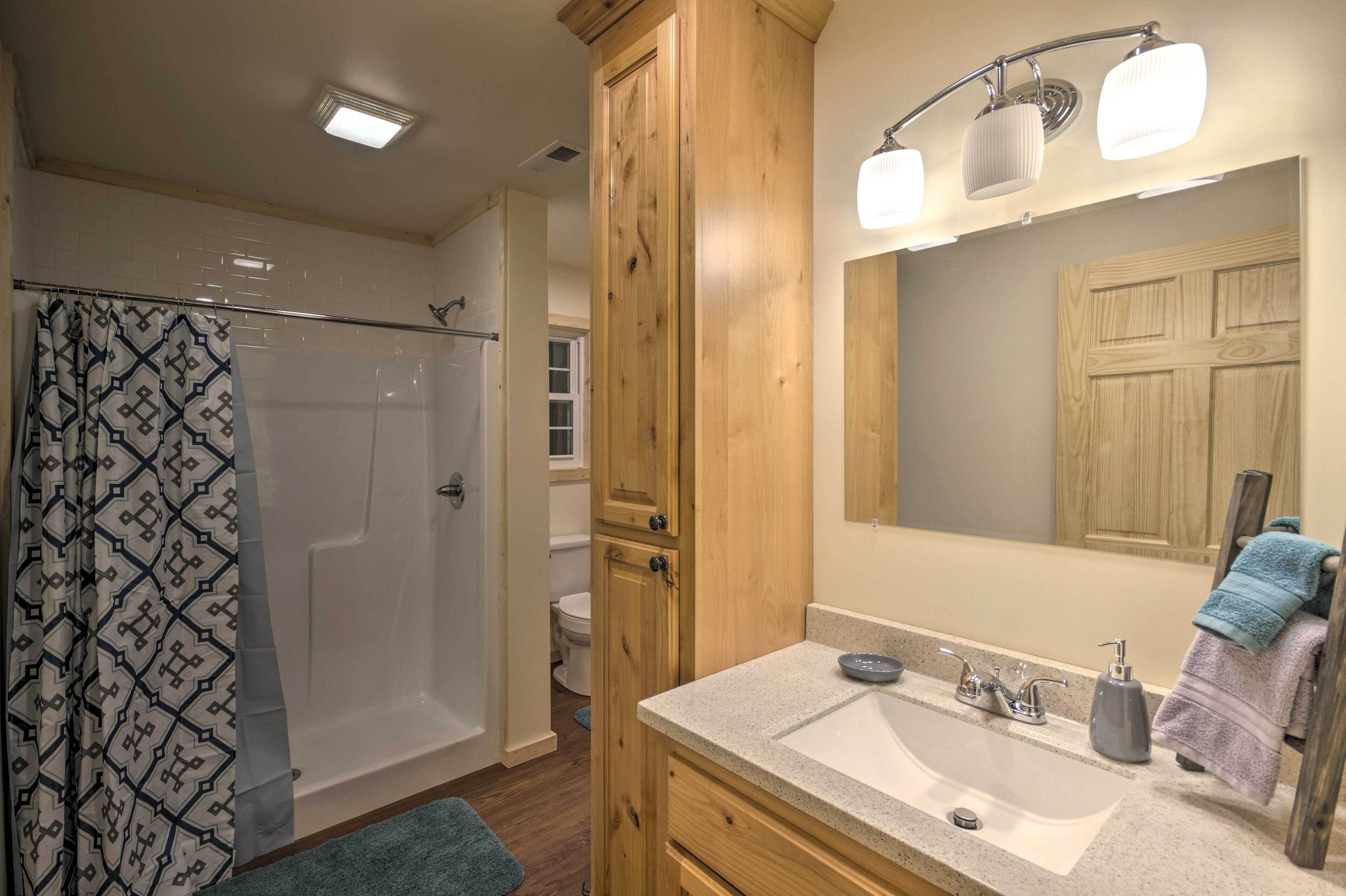 A bathroom with a shower, toilet, wooden cabinet, sink, and a mirror with lights.