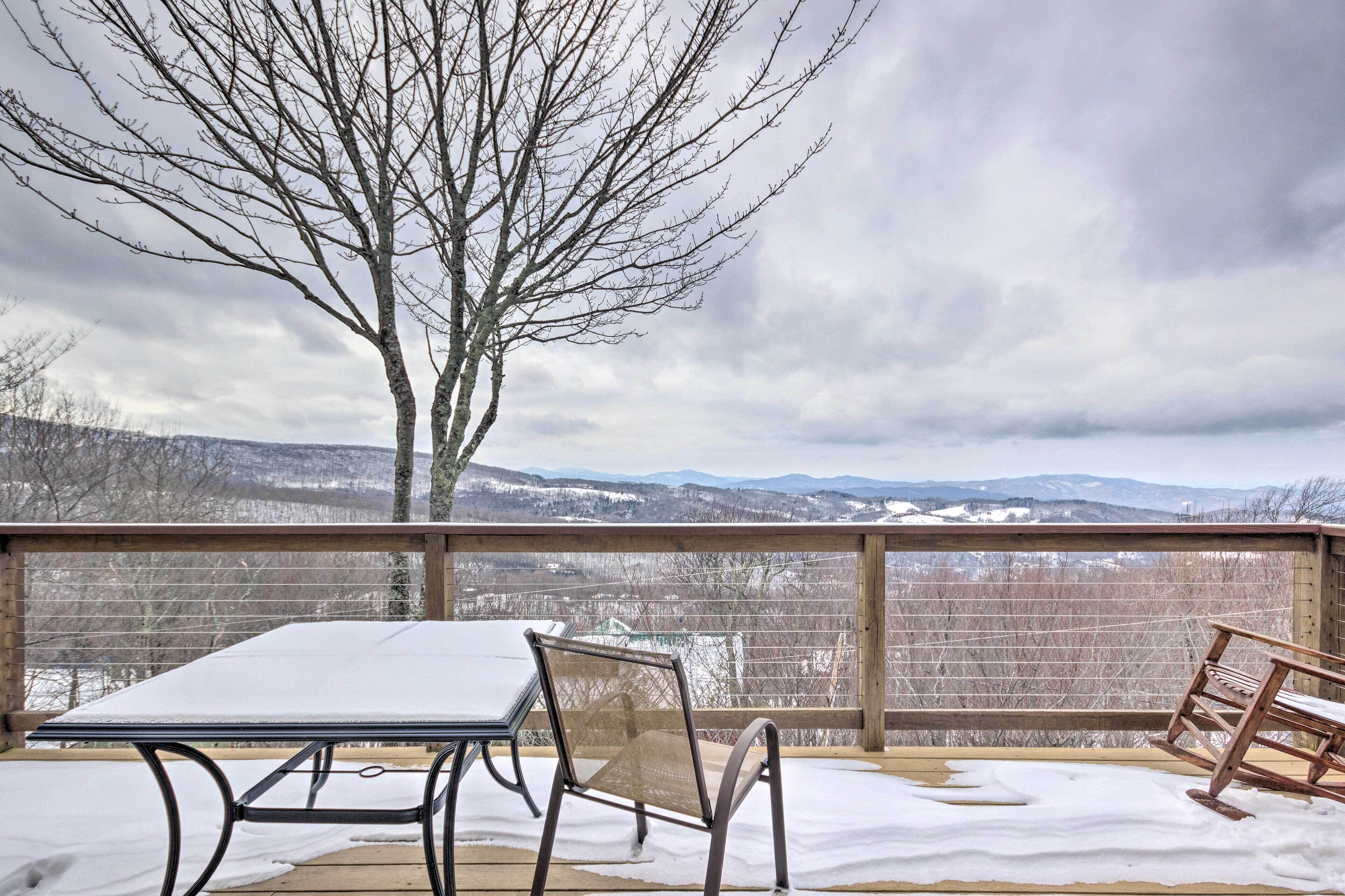 A snow-covered deck with a table and chairs, overlooking a snowy landscape and mountains.