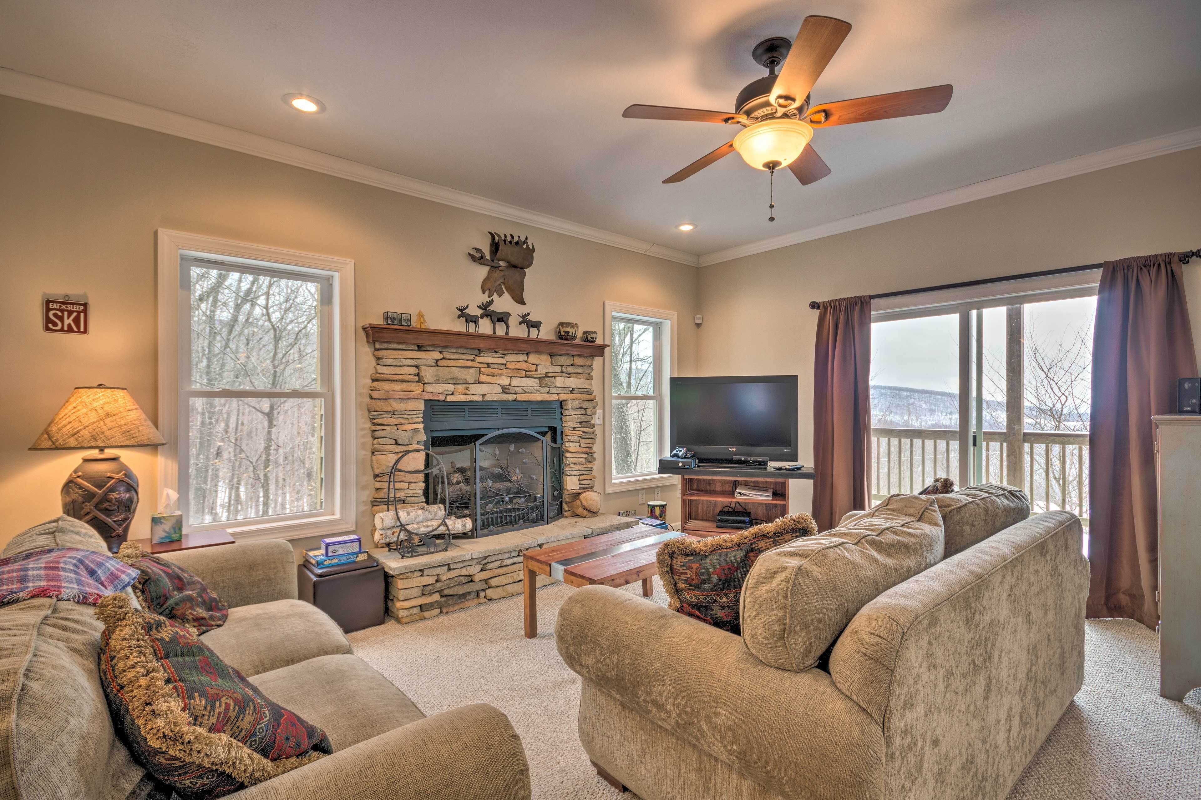 A cozy living room with a stone fireplace, comfortable seating, a flat-screen TV, and large windows with curtains.