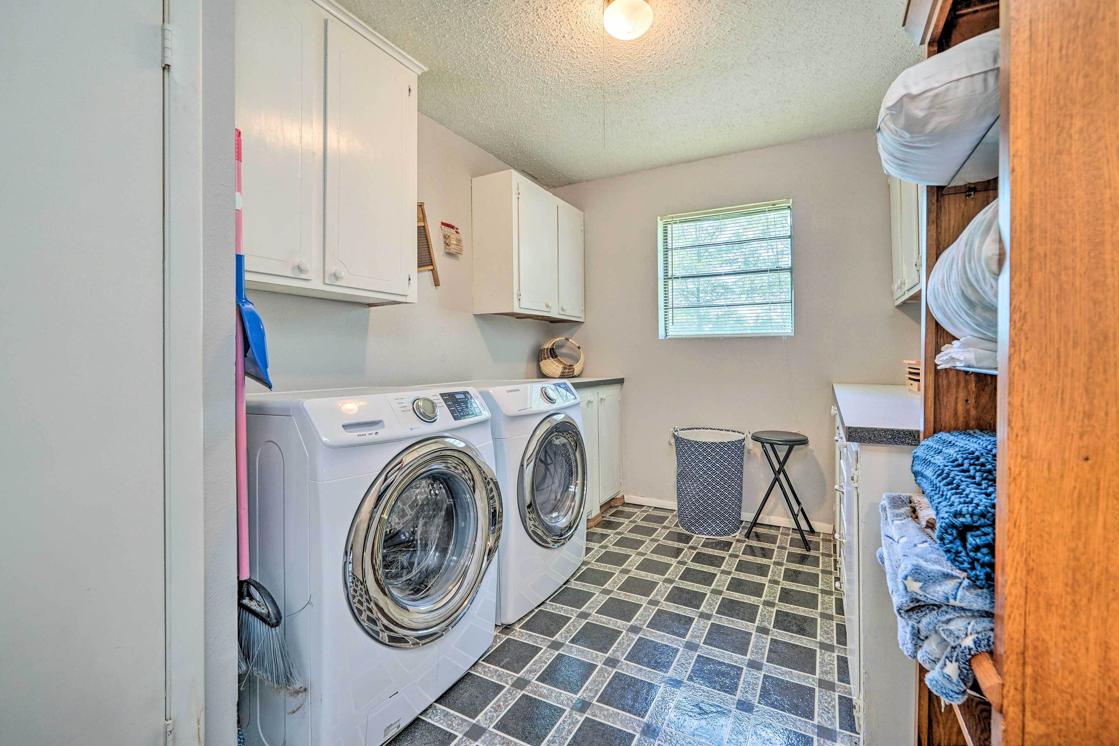 A laundry room with two front-loading washing machines, a window with blinds, a trash can, and a small stool.