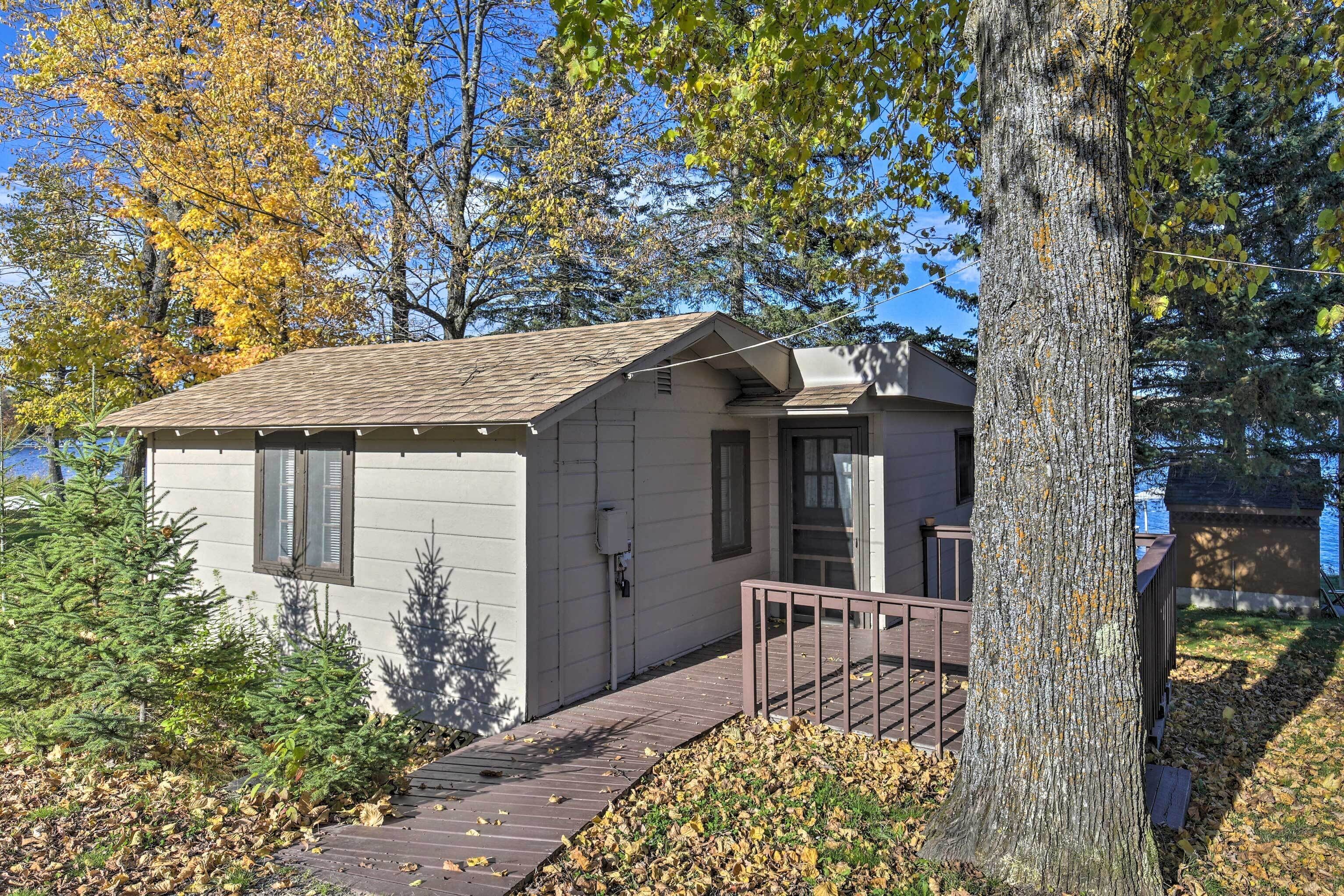 A small cabin with a wooden deck and a metal railing, surrounded by trees and a body of water.