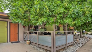 Une petite construction dotée d’un toit en métal et de poutres en bois, entourée d’un chemin de gravier et d’un arbre.