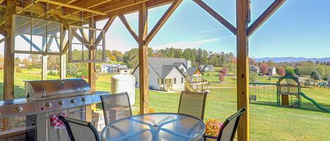 An outdoor patio with a grill, chairs, and a table overlooking a residential area with houses and a playground.
