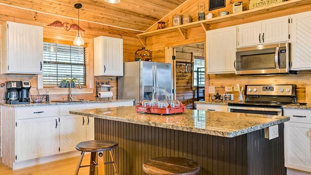 A kitchen with wooden cabinets, a granite countertop, a stainless steel refrigerator, a microwave, and an oven. There are two bar stools and a hanging light fixture. - Cozy New Braunfels Family Cabin w/ Porch & Views! (New Braunfels)