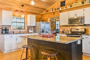 A kitchen with wooden cabinets, a granite countertop, a stainless steel refrigerator, a microwave, and an oven. There are two bar stools and a hanging light fixture.