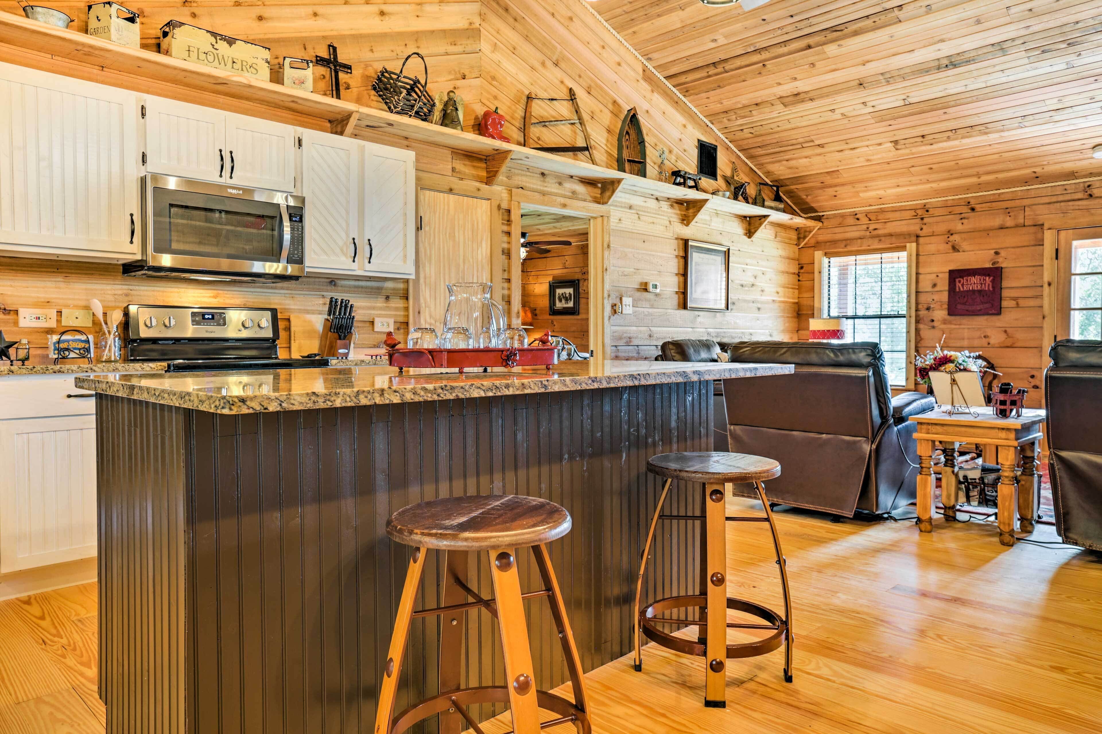Interior de una cabaña de madera con cocina que incluye un microondas de acero inoxidable, horno empotrado y encimera de granito. Hay dos taburetes de madera y una pequeña mesa con una guitarra.