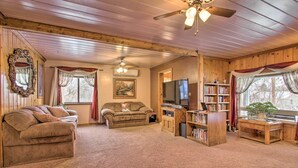 A cozy living room with a wooden ceiling, a ceiling fan, a television, a bookshelf, and a large window with curtains. - Cozy Home w/ Hot Tub: Half Mi to Salmon River! (Salmon)