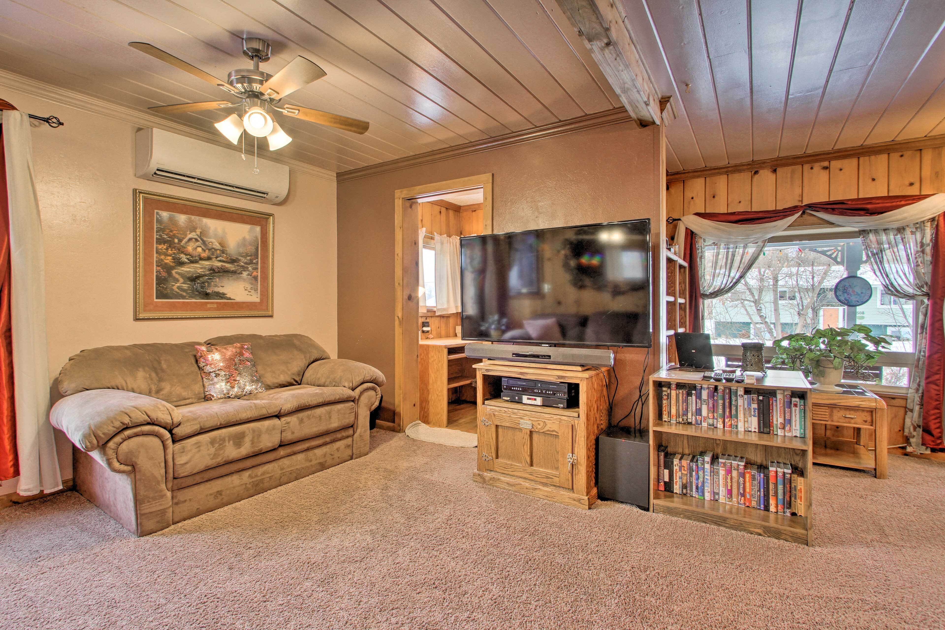 A cozy living room with a ceiling fan, a flat-screen TV, a bookshelf, and a window with curtains.