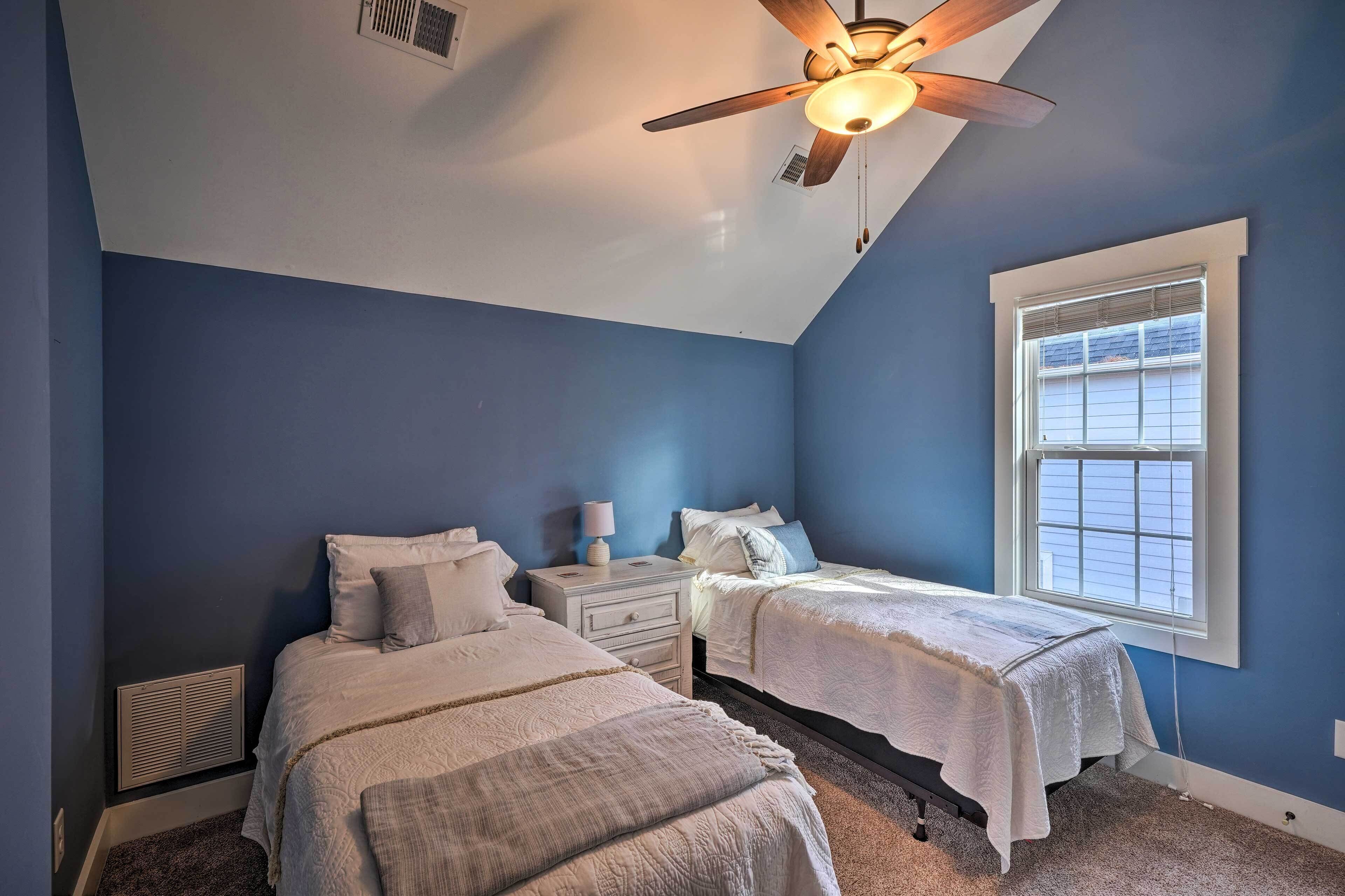 A bedroom with two beds, a ceiling fan, a window with blinds, and a nightstand with a lamp.