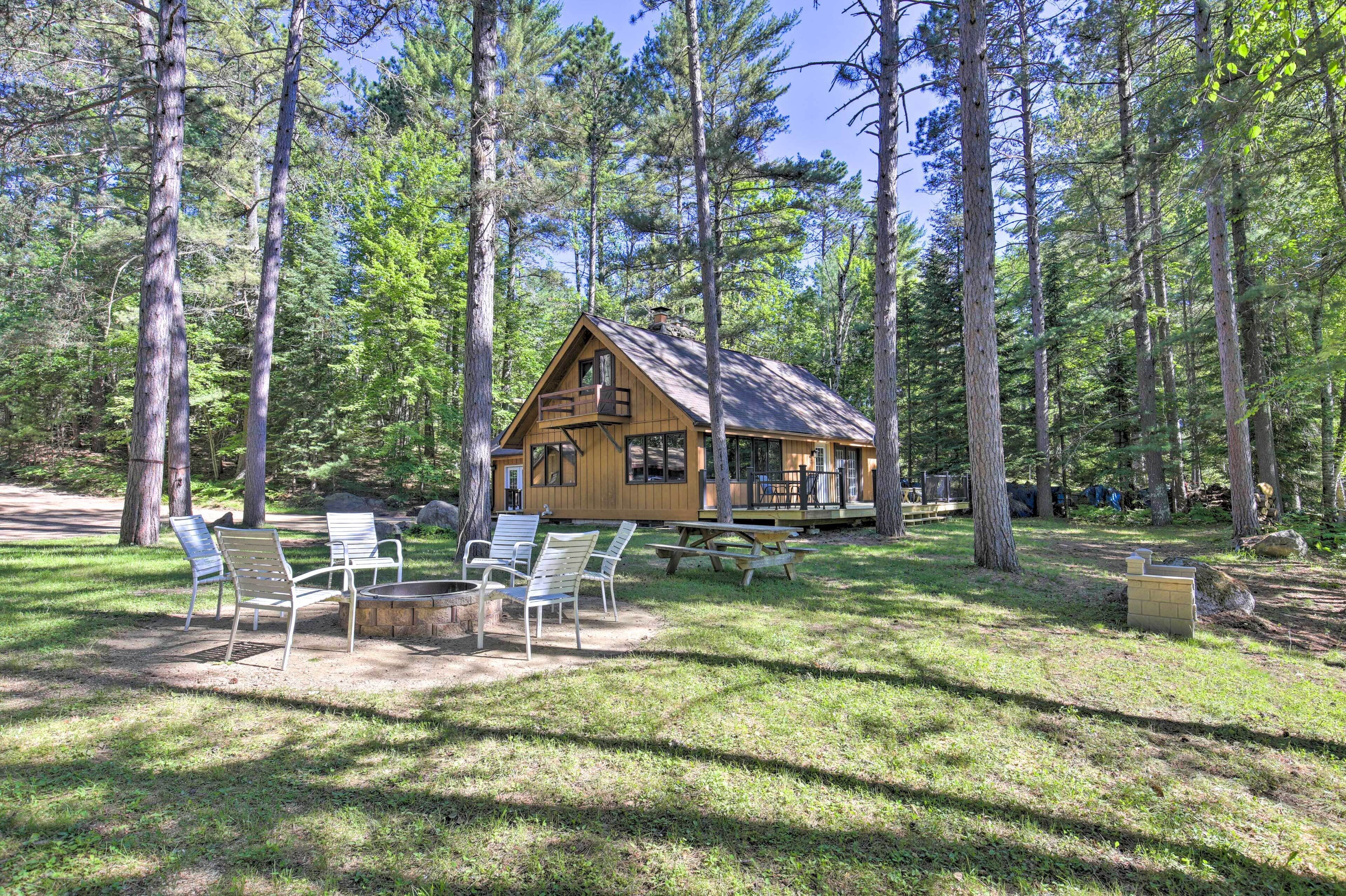 A cabin with a deck, surrounded by a forest. There are picnic tables and chairs in the clearing.