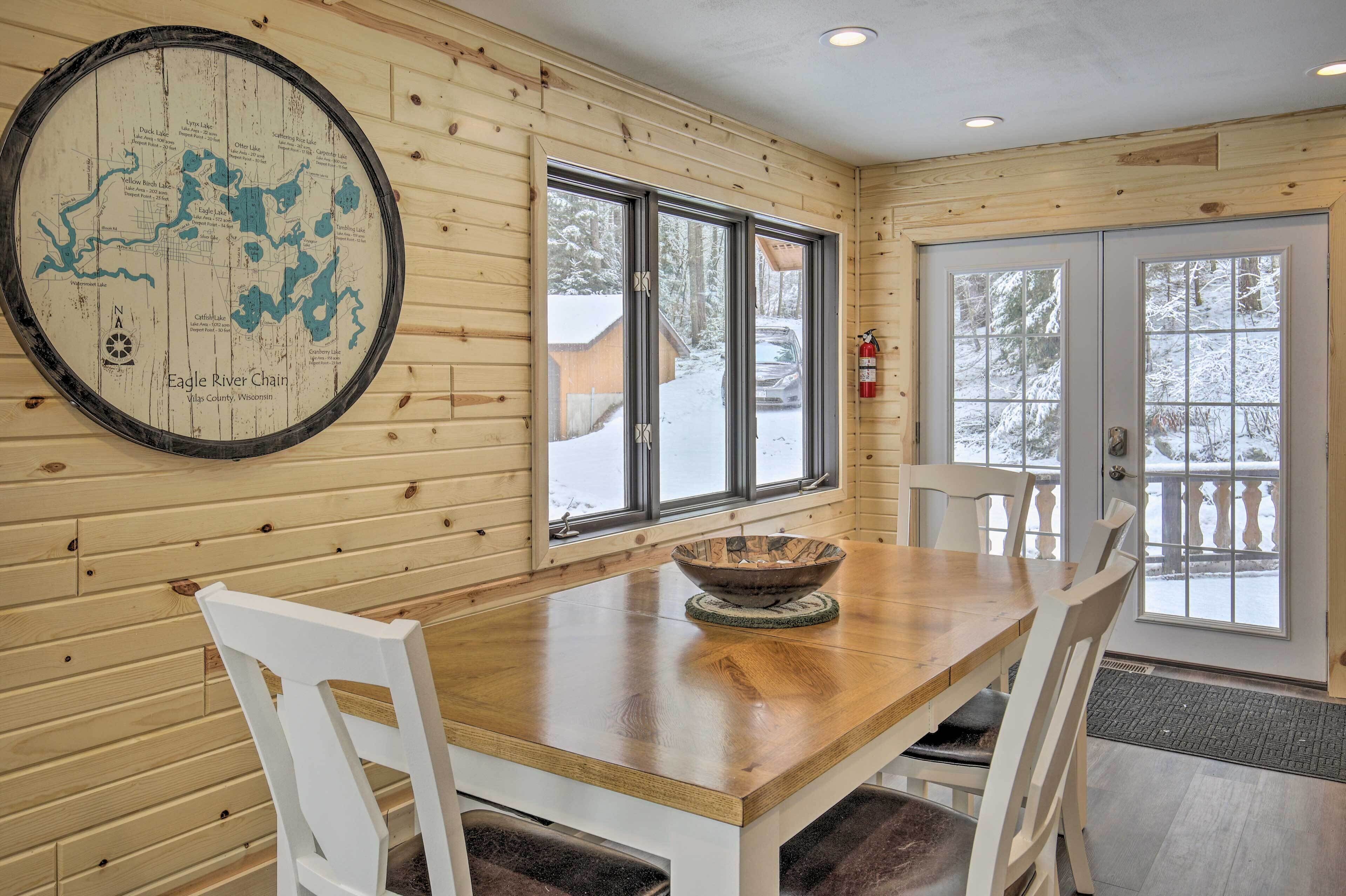 A wooden interior with a map on the wall, a wooden table with chairs, and a view of a snowy landscape through large windows.