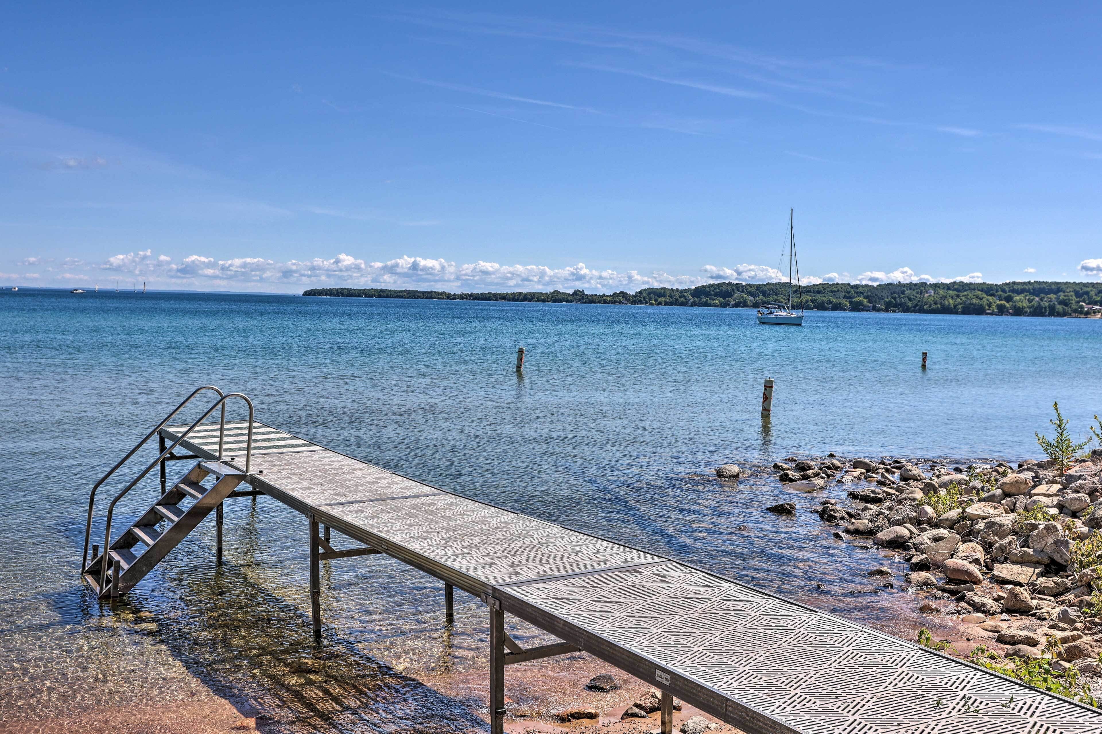 A clear waterfront with a dock extending into calm water, a sailboat in the distance, and a rocky shoreline.