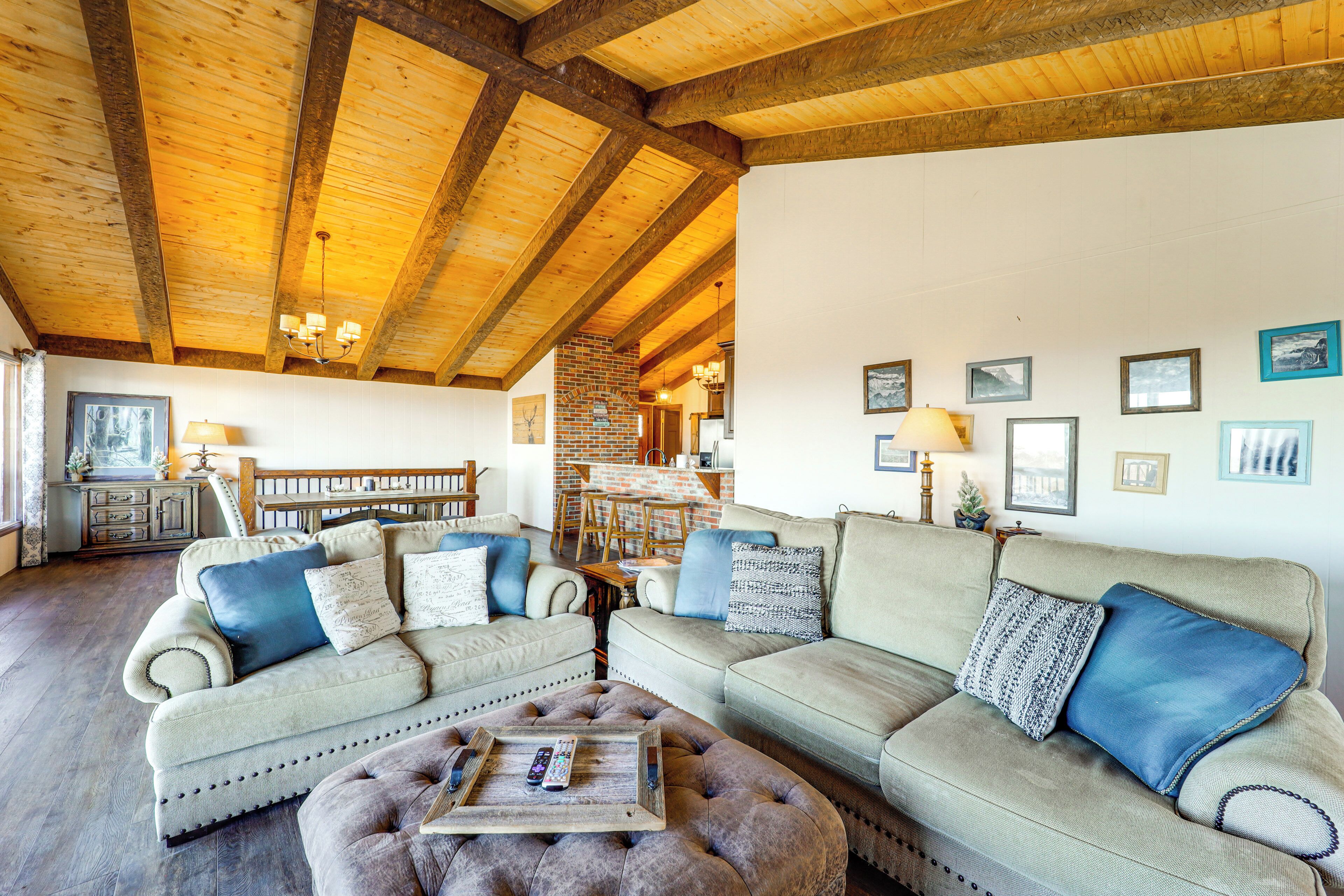 Una sala de estar acogedora con un techo alto de madera, un sofá beige, un otoman redondo y un comedor al fondo.