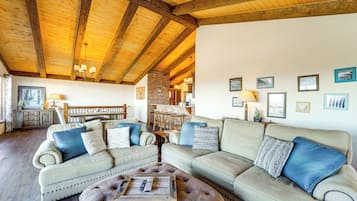 Una sala de estar acogedora con un techo alto de madera, un sofá beige, un otoman redondo y un comedor al fondo.