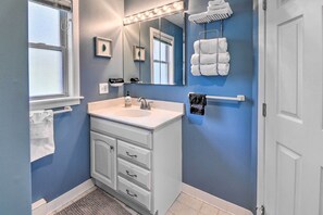 A bathroom with a white vanity, a large mirror with lights, a towel rack, and a door.