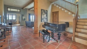 A spacious living room with a grand piano, a staircase, and a dining area with a view of the outdoors. - Charming Mountain Estate w/ Views + Fire Pit! (Cerrillos)