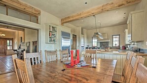 A spacious dining area with a large wooden table and chairs, a kitchen in the background, and a high ceiling with exposed beams. - Charming Mountain Estate w/ Views + Fire Pit! (Cerrillos)
