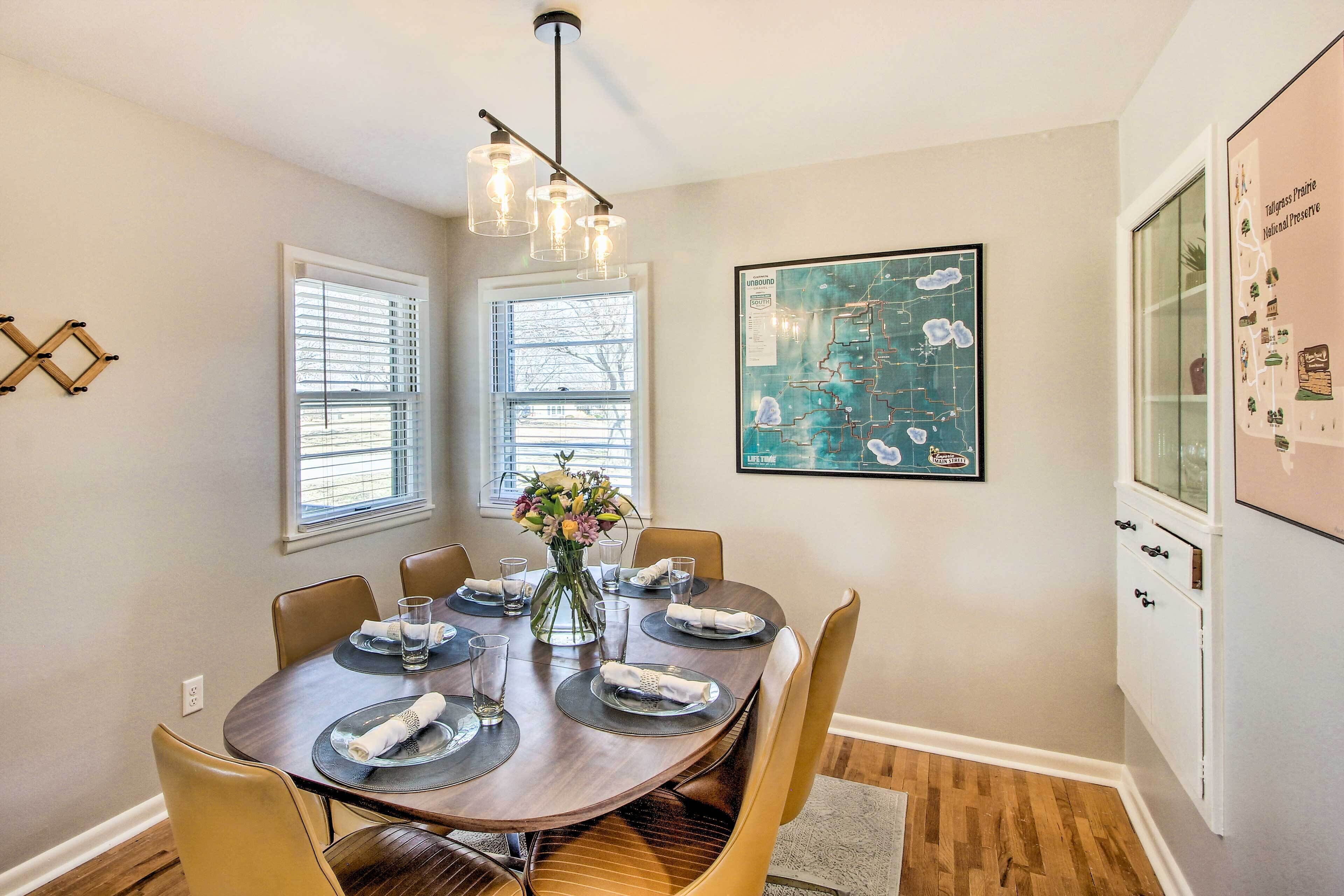 A dining area with a round table set for six, a vase with flowers, and a map on the wall.
