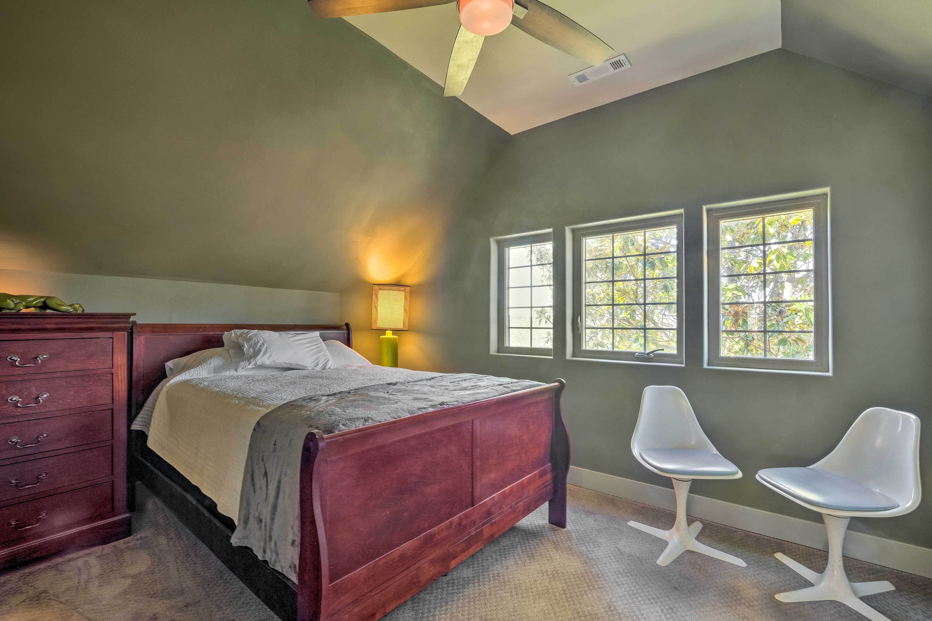 A bedroom with a wooden bed, a dresser, two white chairs, and a window with a view of trees.